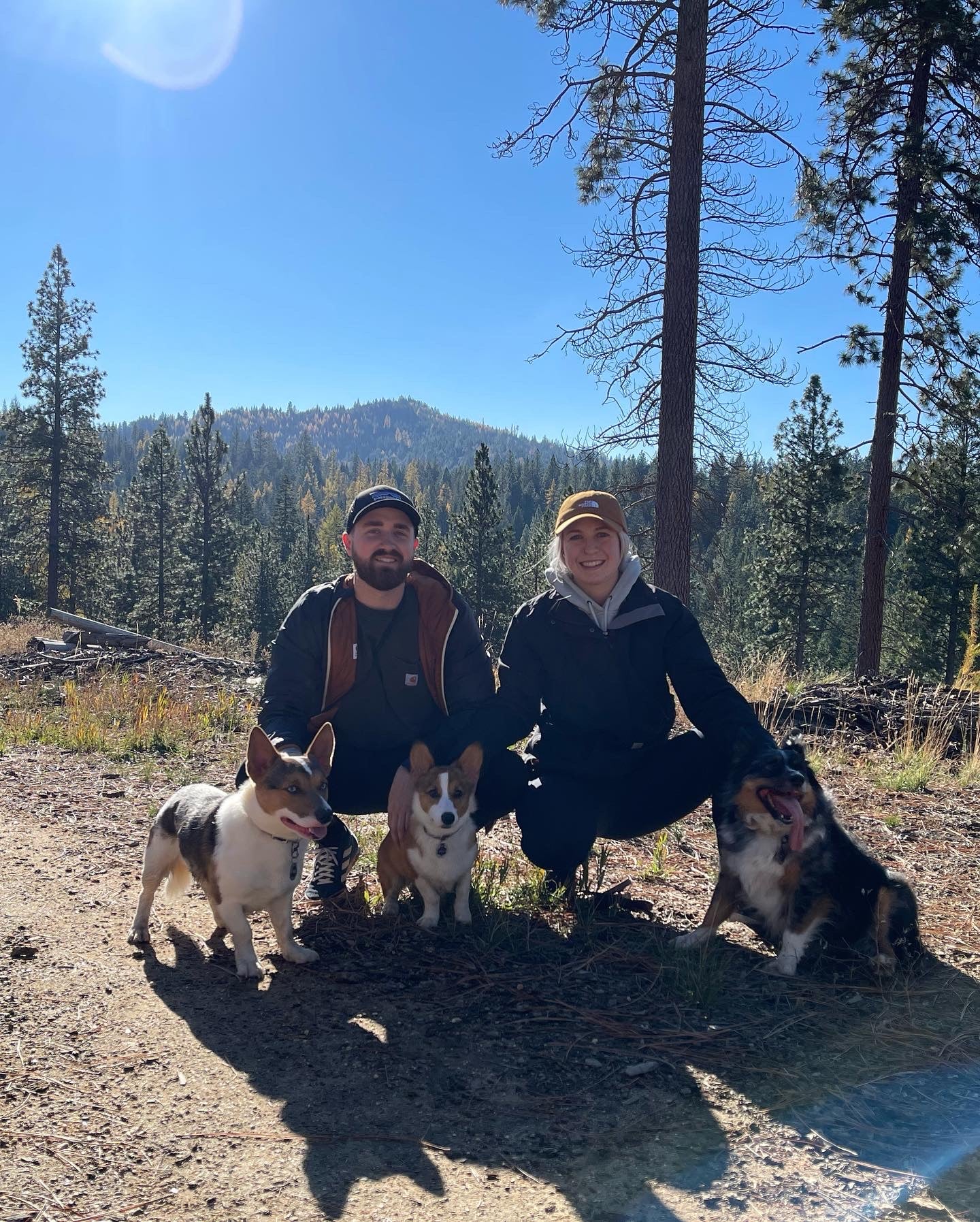 Two people and three dogs posing on a trail with trees and mountains in the background