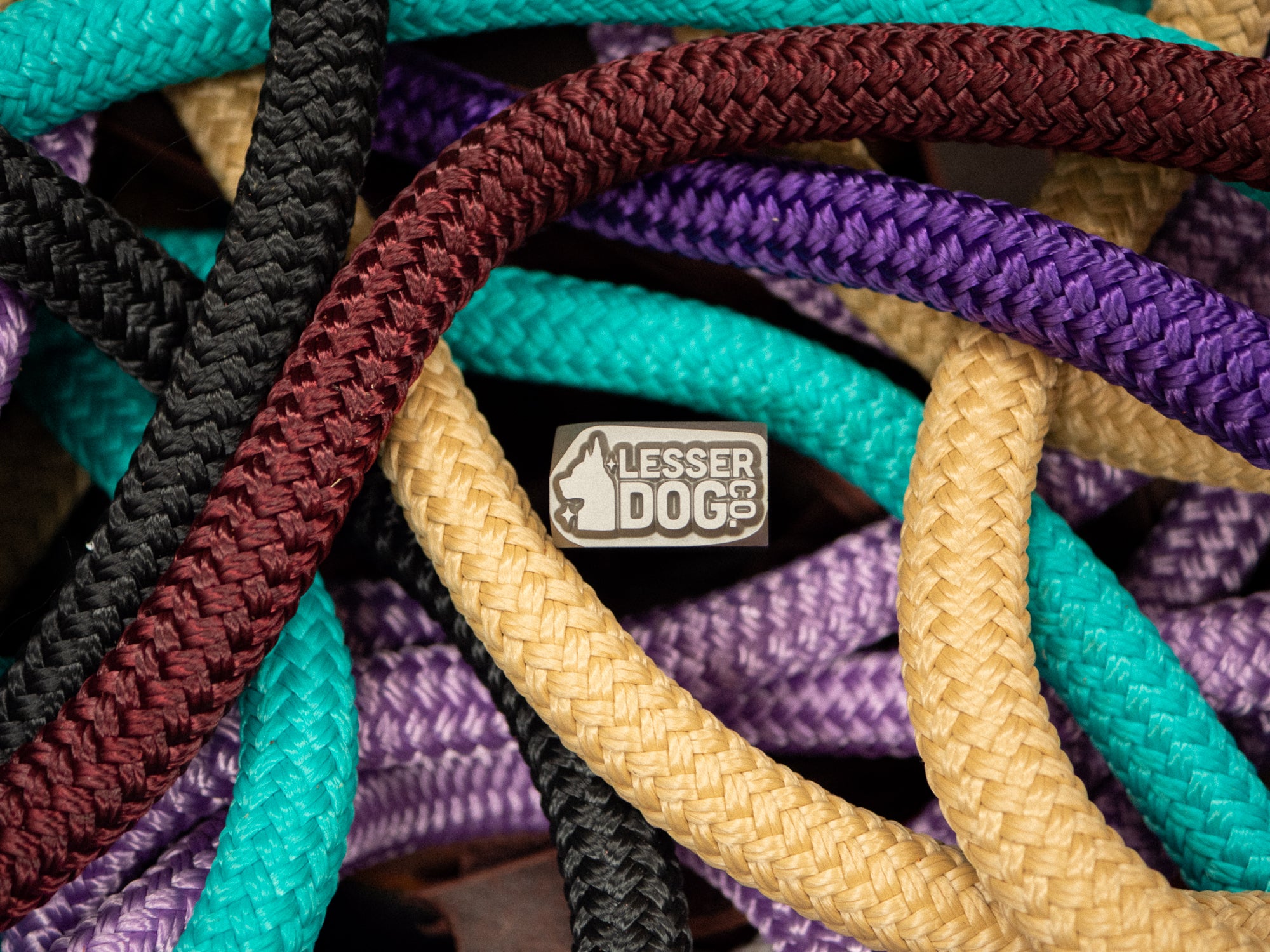 A close-up of braided rope cords in multiple colors, including teal, purple, beige, black, and burgundy, with a small metal badge reading "Lesser Dog Co" visible near the center.
