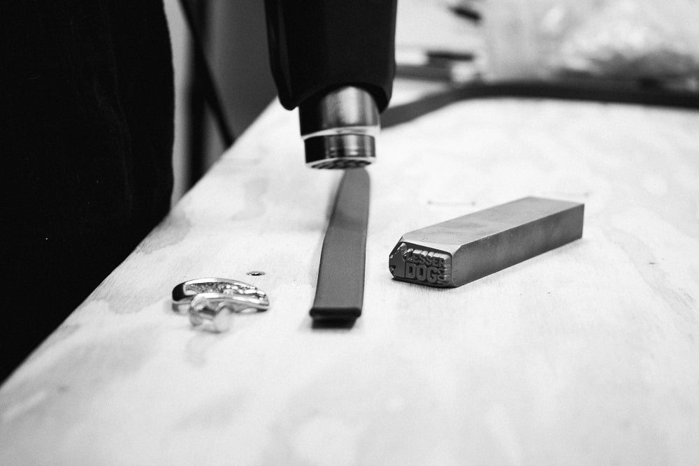 A close-up black and white photo of a metalworking setup on a workbench, showing a heat gun above a metal bar, a small metal ring, and a rectangular metal block with the brand name 'DOG' visible on it.