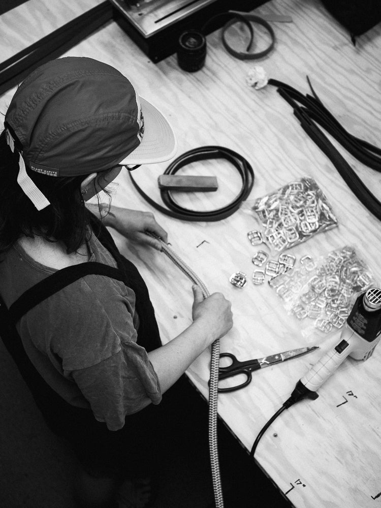 A black and white overhead shot of a person wearing a cap and apron, working at a wooden table with jewelry-making tools, wire, beads, and scissors.