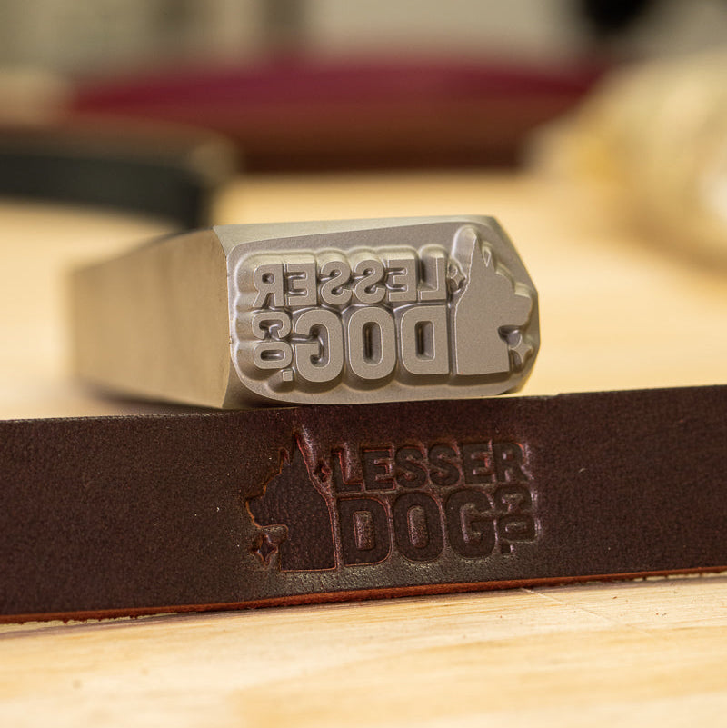 A silver metal dog-shaped stamp with the words 'LESSER DOG' engraved on the face, resting on a dark brown leather strap that is also embossed with the same logo, set against a warm, blurred background.